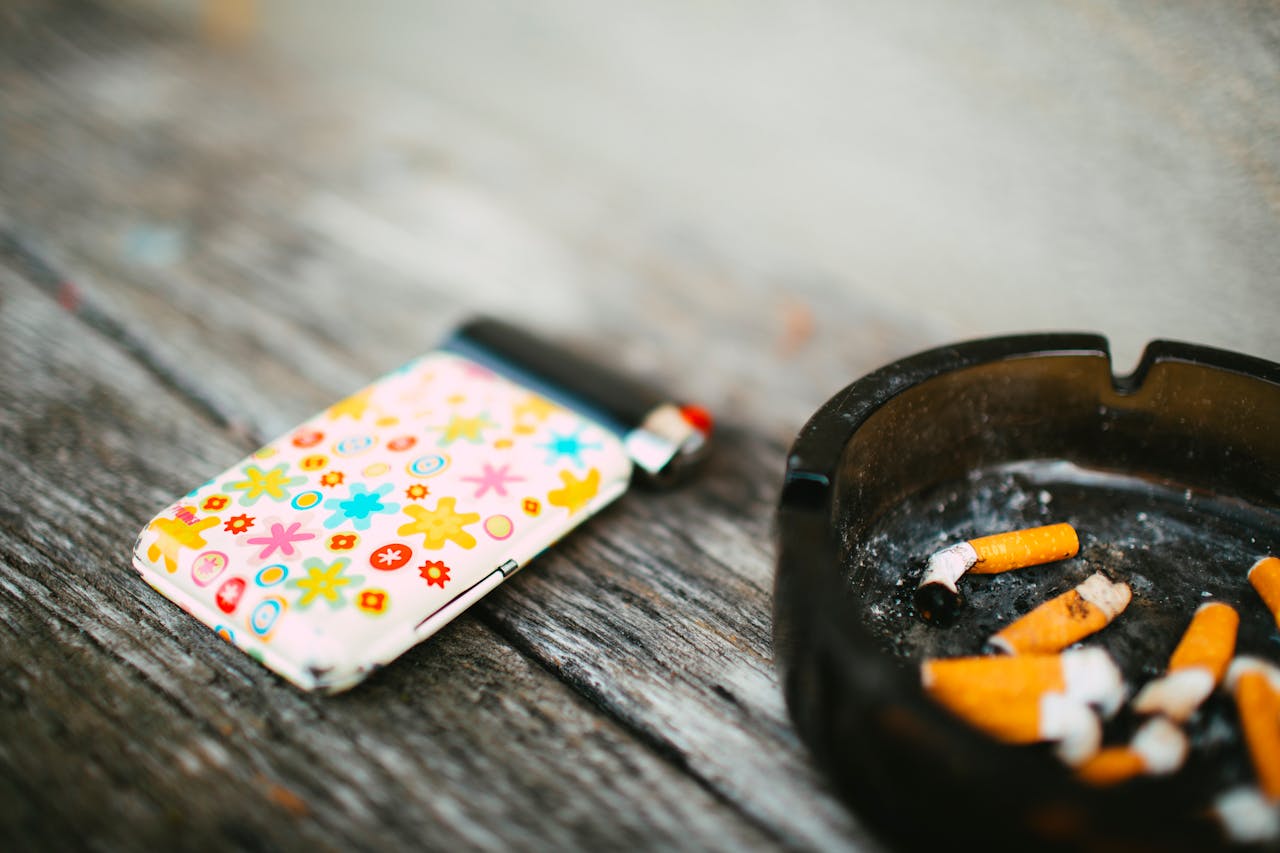 A vibrant lighter sits next to a black ashtray filled with cigarette butts on a wooden surface.